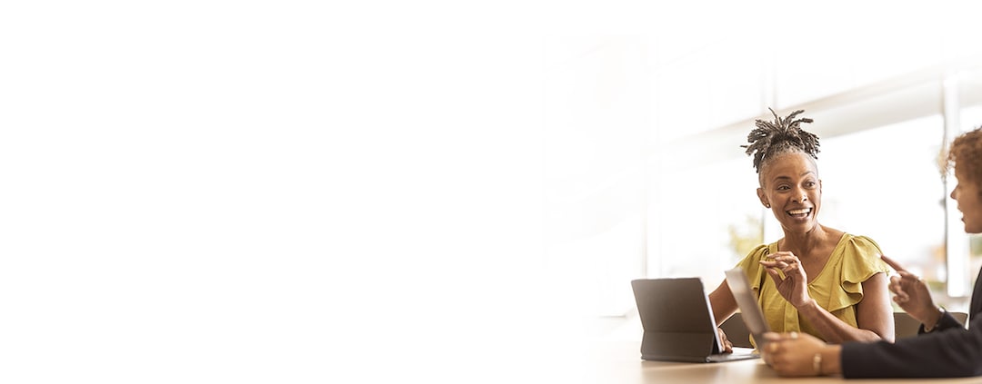 An image of two women sitting at a table. The focus is on a woman in front of a laptop, smiling and making a hand gesture. Next to her is another woman wearing a suit jacket, holding a tablet, pointing and speaking.