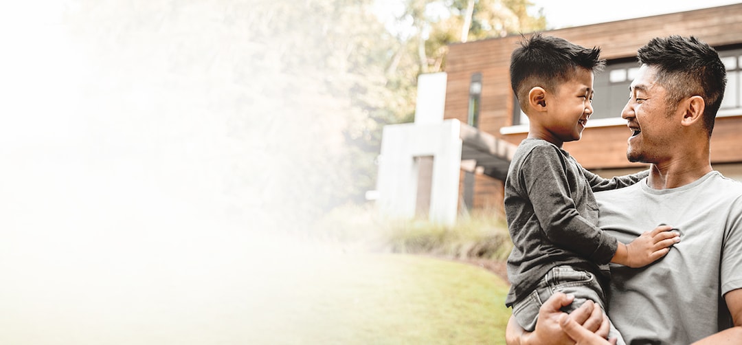 Young man, a father, carrying and smiling at young boy, his son, who is smiling back. Modern home in the background.