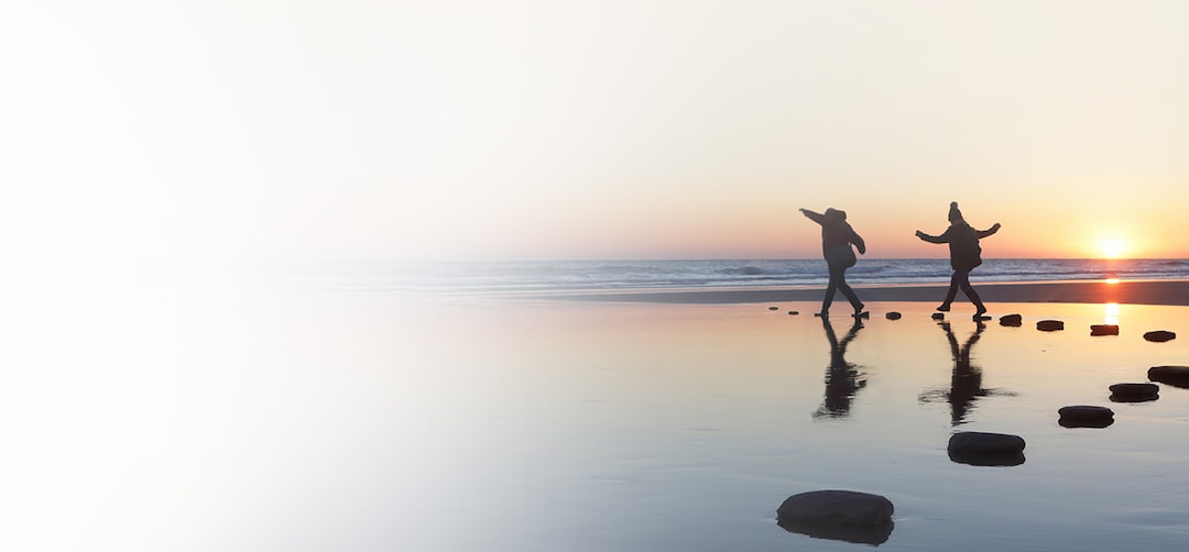 An image of a beach at sunset with 2 people skipping at the coastline on the beach stones.