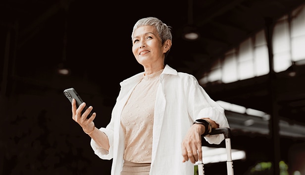 A woman with short gray hair standing indoors with a smartphone and suitcase in an industrial-style setting.