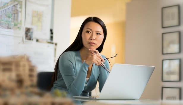 An image of an Asian woman in front of her open laptop, holding her eyeglasses and looking to her right with a serious gaze.